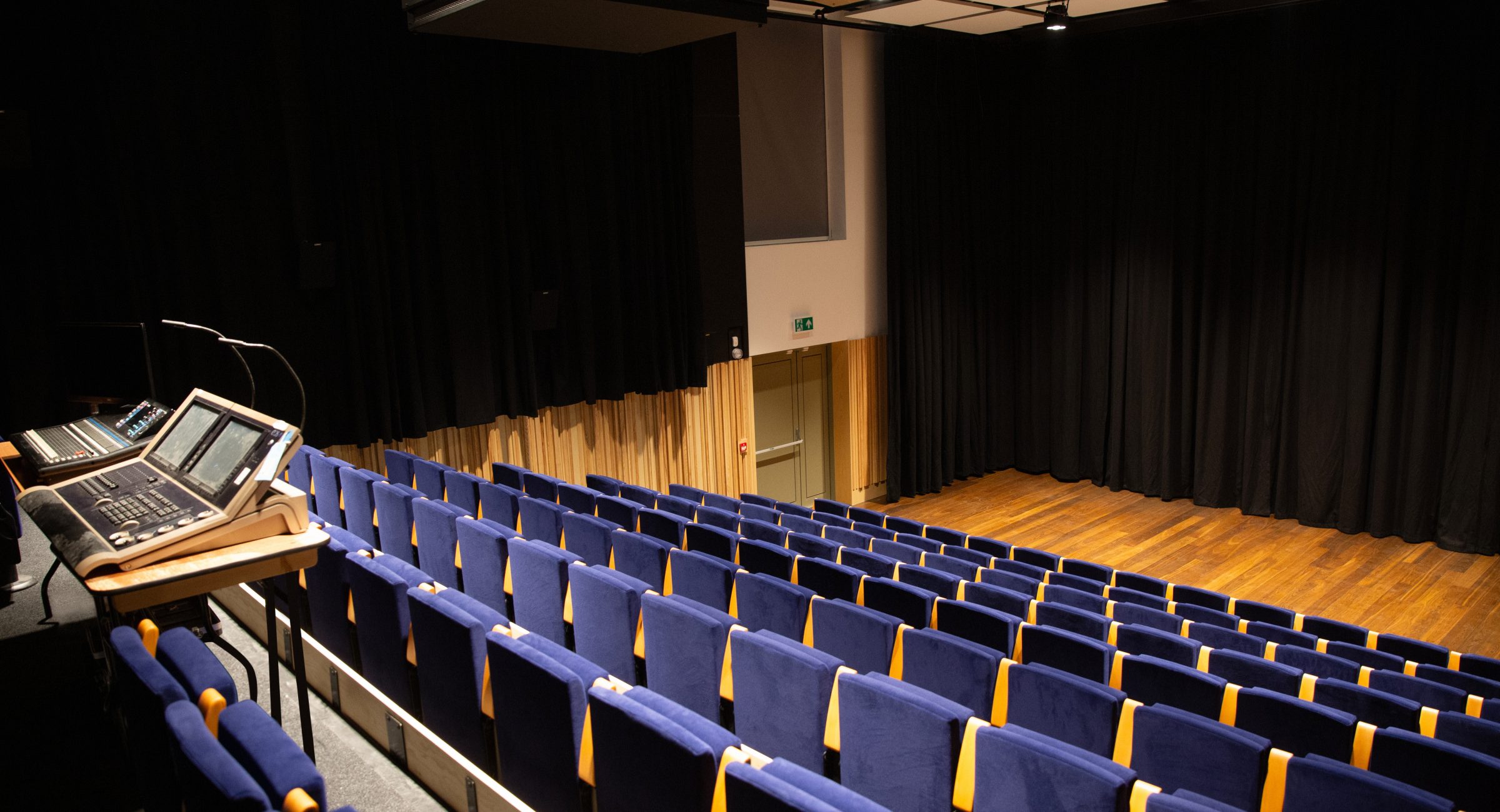 A black box style room with navy blue chairs and a lighting desk with technical equipment at the back.