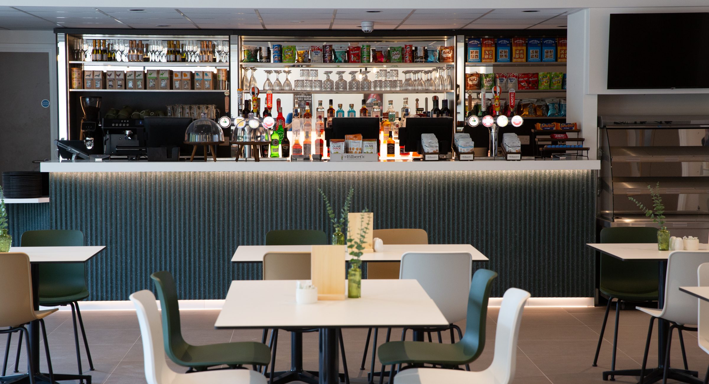 Brightly dressed full stocked blue bar with displays of items and bottles in the background, with cafe tables and chairs in the foreground.