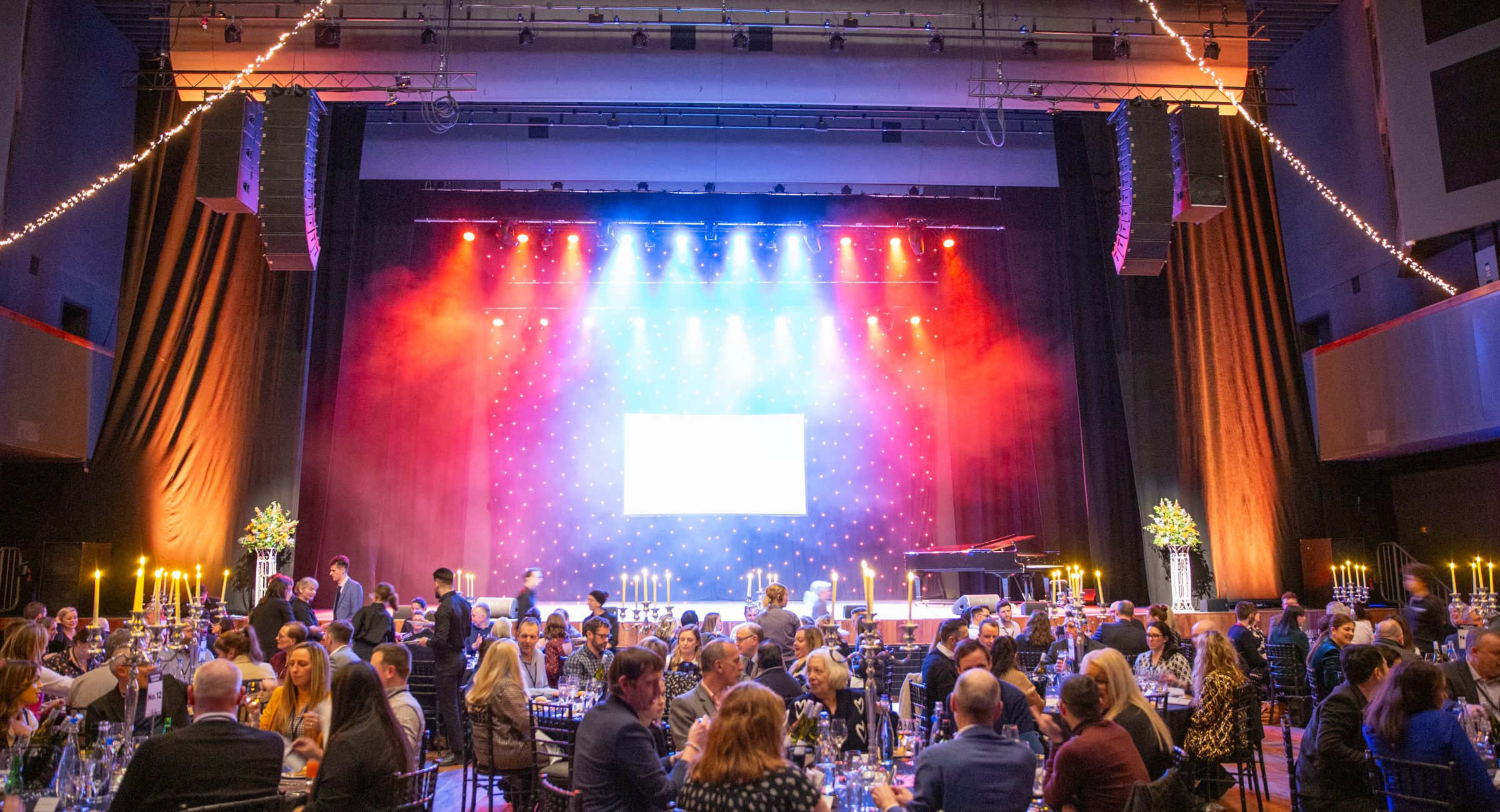 A red and blue lit stage with a screen for presentations and star backdrop, guests sat for a banquet waiting to be served, with festoon lights at the edge of the image.