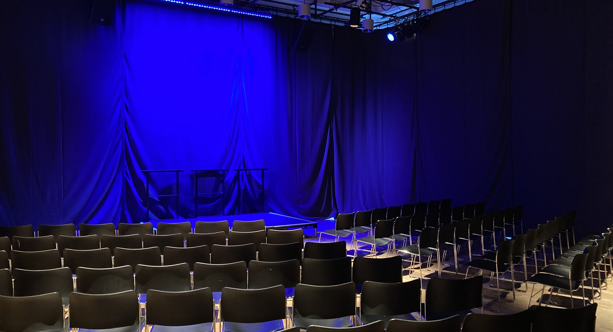 Six rows of black chairs in front of a blue lit black curtained stage space.
