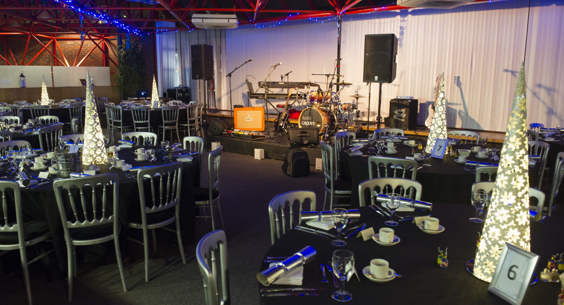A shot of tables dressed with white lit Christmas trees with a stage in the background set up for a band with a drum set and speakers.