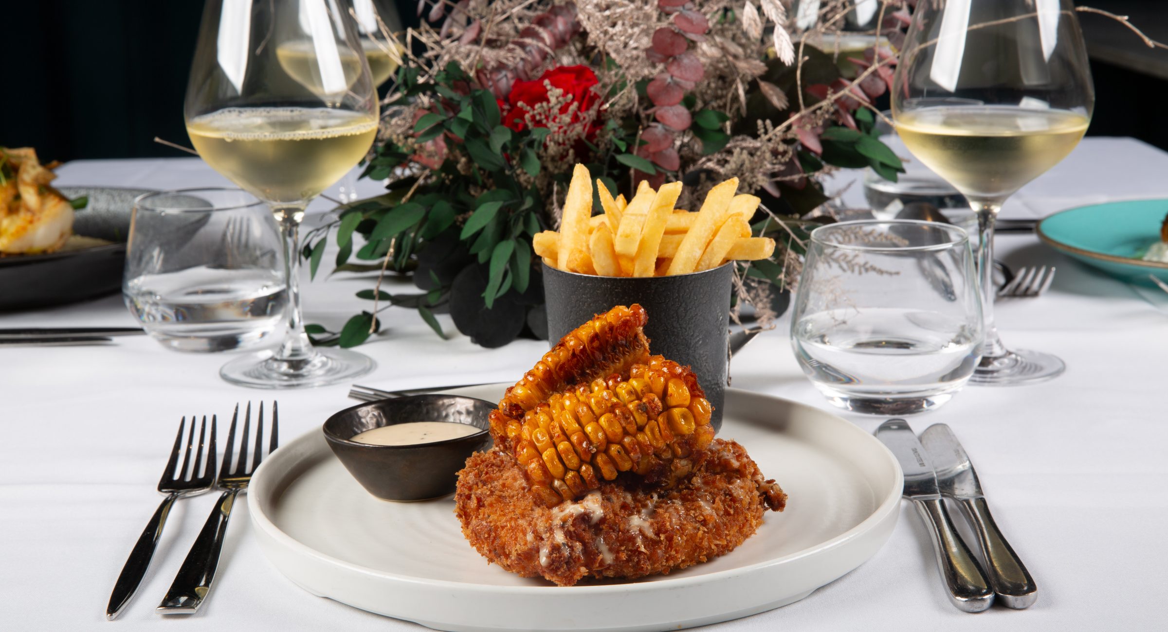 A table featuring a delicious plate of elevated chicken and sweetcorn ribs and a pot of french fries. A beautiful floral centerpiece sits behind this, with glasses of wine, water glasses and cutlery.