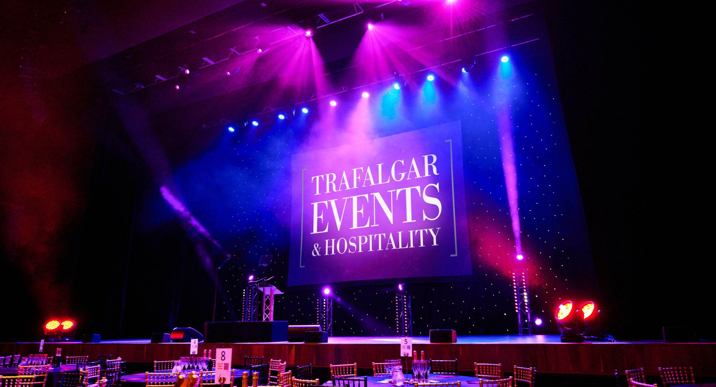A room lit in pink and blue with a screen onstage displaying Trafalgar Events & Hospitality and tables with silver chairs around them.