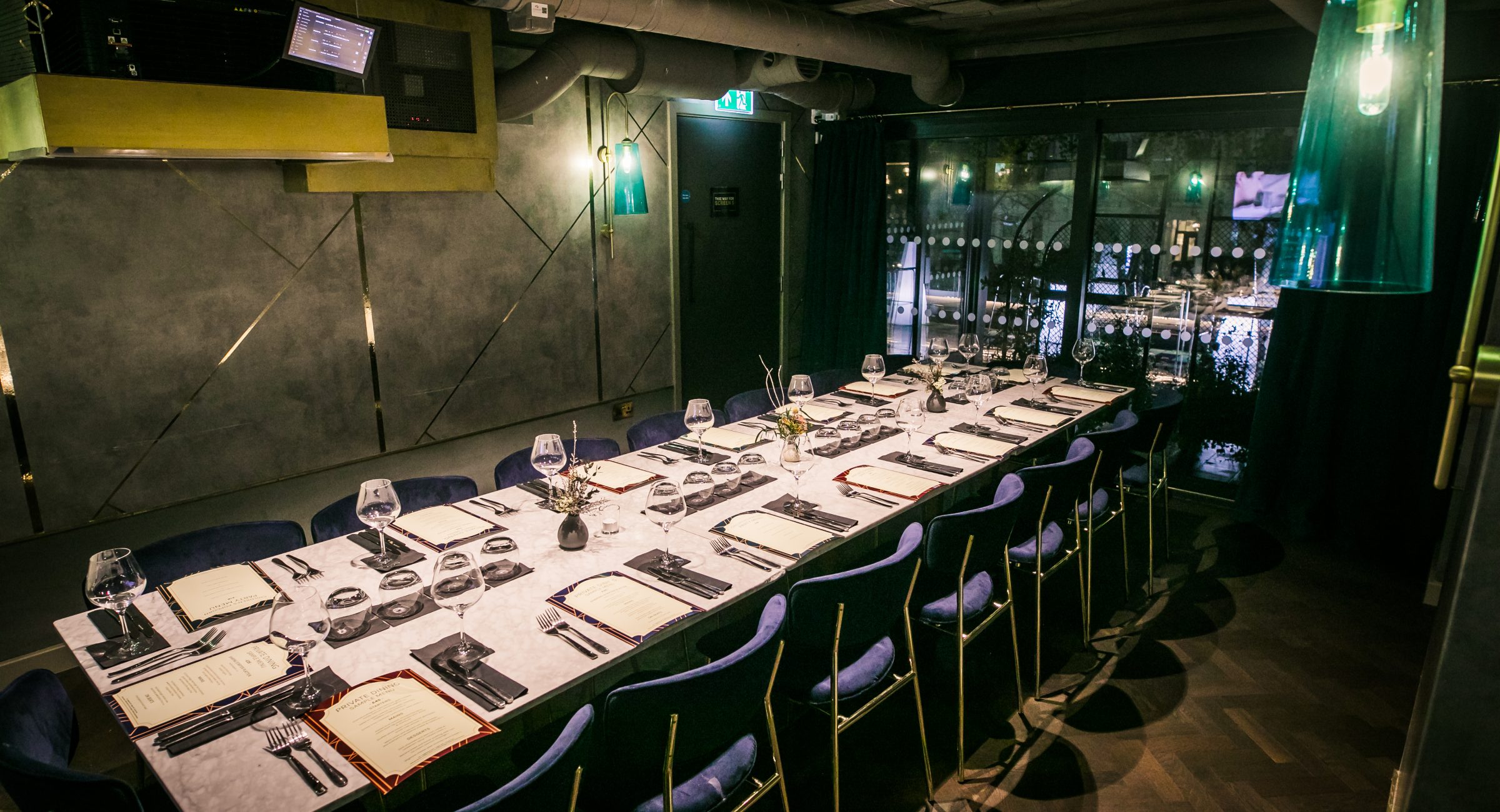 A long marble dining room table with blue chairs around it in the centre of a room with marble style wall paper and projector visible.