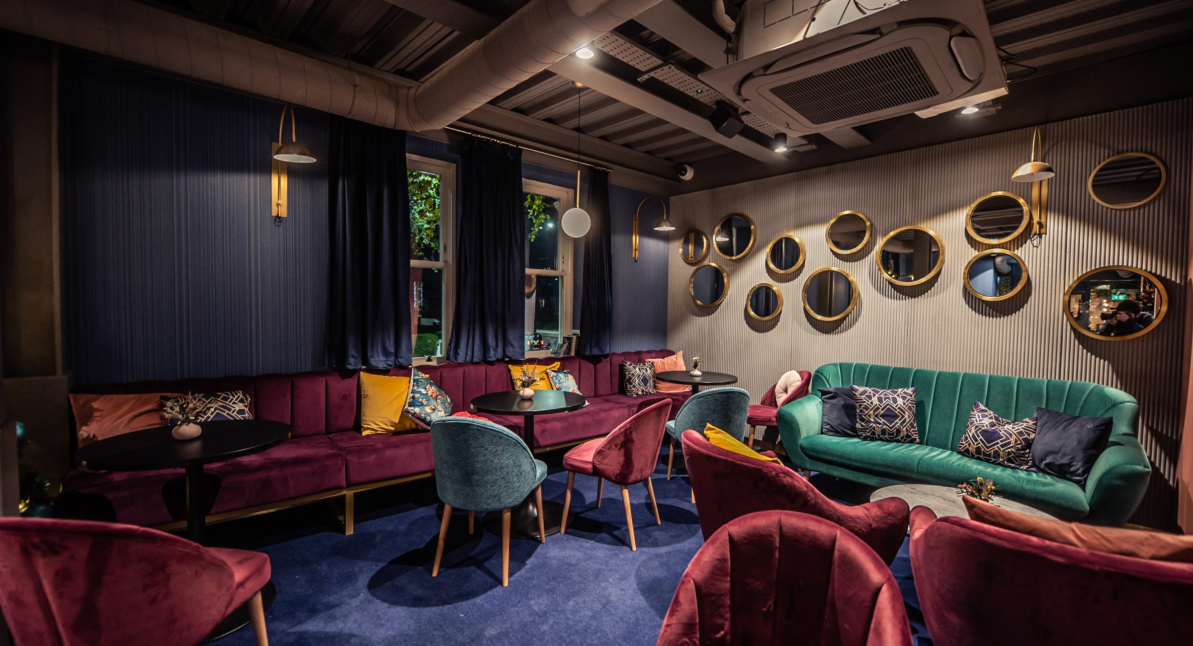 Brightly coloured empty sofas and chairs on a navy blue carpet and circular gold mirrors on a textured wall.