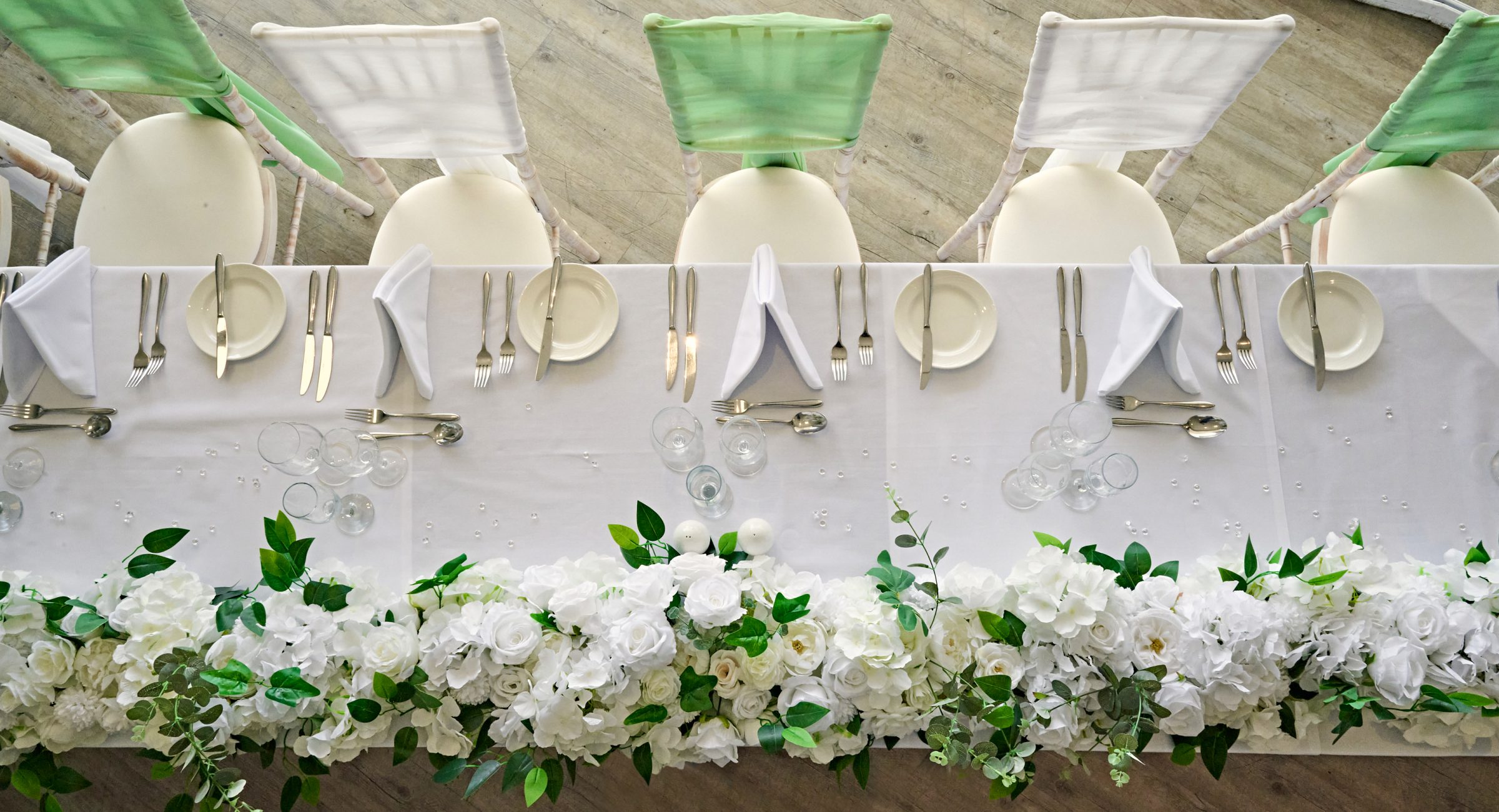 A vertical view of the top table of a wedding; dressed with green foliage and white flowers, green and white sashes on the chairs and laid up with cutlery, side plates and cloth napkins.