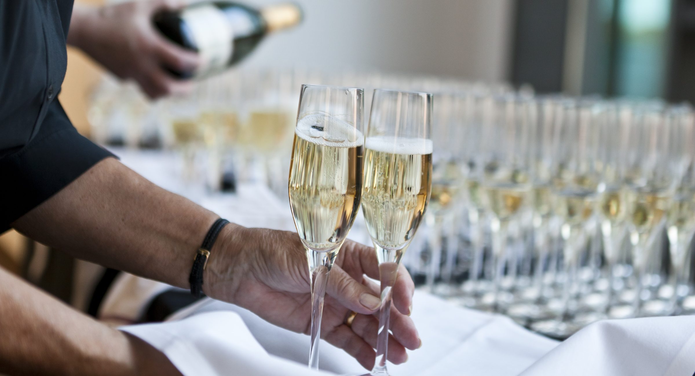 Bubbling glasses of champagne, with two glasses on a tray in the foreground and more glasses in rows being topped up with a bottle in the background.